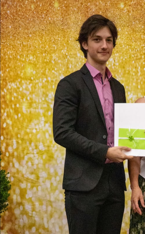 Junger Mann in Anzug und rosa Hemd hält ein weißes Buch mit grünem Band vor goldenem Hintergrund.
