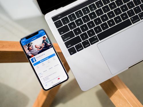 Smartphone mit Facebook-App neben einem geöffneten Laptop auf einem Holztisch.